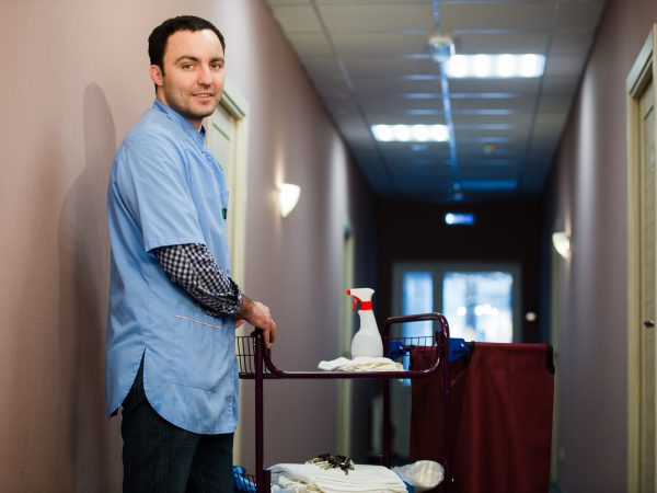 A man who is on the hotel cleaning crew staff is smiling with a towel, pillow and vacuum in the process of cleaning the hotel rooms and delivering top-knotch service to the guests